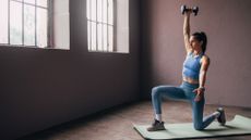 A woman kneels on a yoga mat in an empty room, with one leg bent before her and the other bent behind her. She also holds a dumbbell in her right hand high above her head.