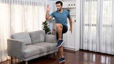 man in a living room setting standing on one foot with the other knee raised to hip height like a standing march. there's a grey sofa to the side of him and windows behind. 
