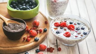 A bowl of white chia seed pudding, with berries, sits on a table next to a chopping board that has another bowl of chia seeds on it and a small bowl of blueberries.