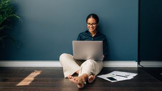 Woman sits on floor with back against a wall and laptop on her lap