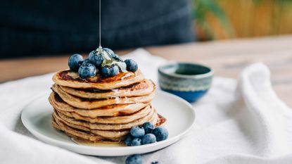 A stack of pancakes sits on a white plate topped with blueberries. Honey is being poured over the pancakes from above.