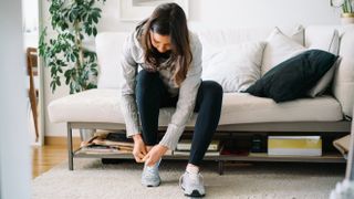 A woman sits in a couch bending forward to tie her shoes. Behind her we see a plant.