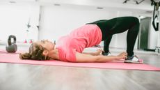 woman on a pink exercise mat in a white space gym background performing a glute bridge, facing away from the camera.