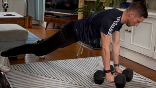 Man in top of a push-up position holding two dumbbells resting on the floor. He is in a domestic setting and wears a blue T-shirt, black trousers and white trainers