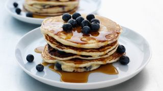 A stack of pancakes on a plate drenched in maple syrup and topped with blueberries.