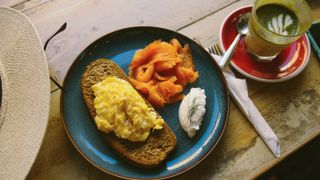 From above, we see a blue plate with a slice of sourdough bread, scrambled eggs and cream cheese. Next to it is a matcha latte.