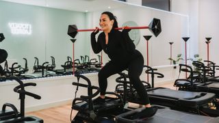 Woman exercising on Lagree Megaformer machine. She holds a light bar across the back of her neck and is squatting with her knees bent and hips pushed back