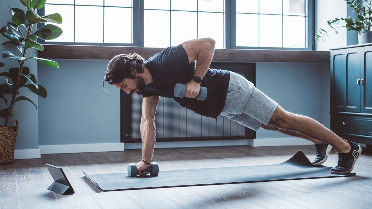 side shot of a man performing a renegade row move with two dumbbells in a living room setting. there's a window behind him and a plant to the side.