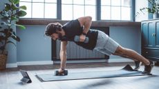 side shot of a man performing a renegade row move with two dumbbells in a living room setting. there's a window behind him and a plant to the side.