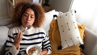 A woman in a stripy jumper eats a bowl of oatmeal. She is on a couch and holds the spoon to her mouth.