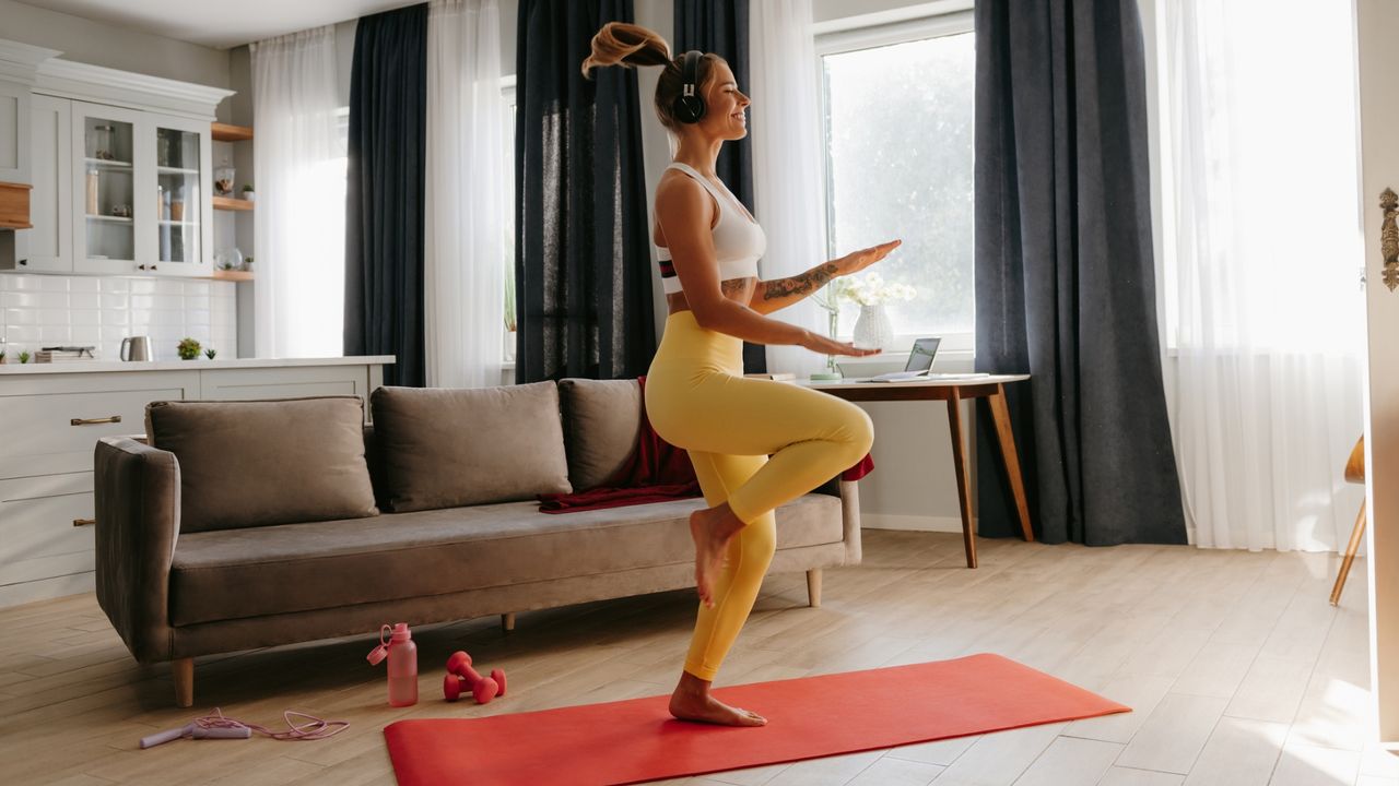 woman in a living room performing a high knees exercise on a red exercise mat. she's facing sideways to the camera wearing yellow leggings and a white top with headphones on and smiling.