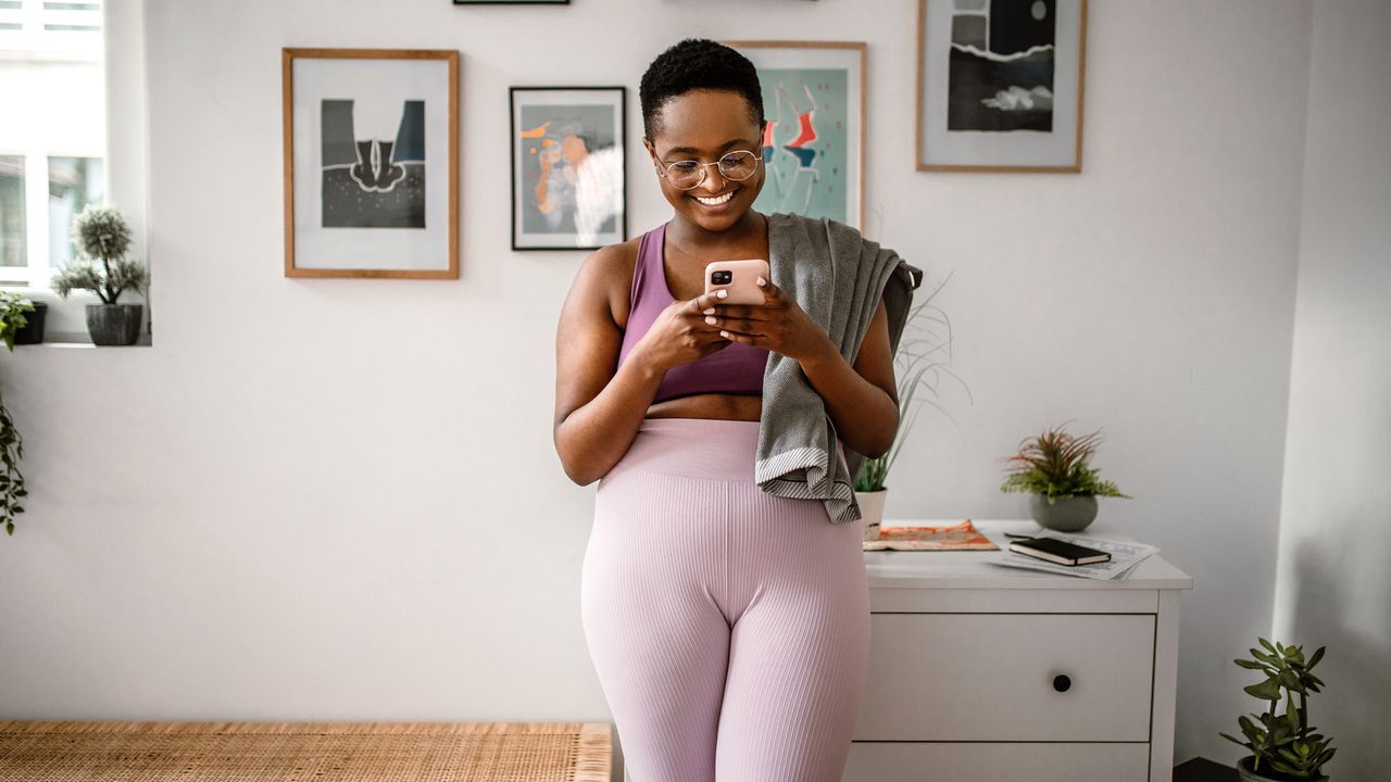 Woman in sportswear leaning against chest of drawers looking at phone with small towel draped over one shoulder