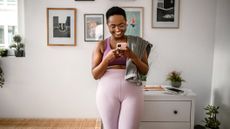 Woman in sportswear leaning against chest of drawers looking at phone with small towel draped over one shoulder