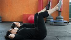 two women dressed in black workout clothes lying on their backs with knees up and bent at 90 degrees about to perform a core strengthening move. 