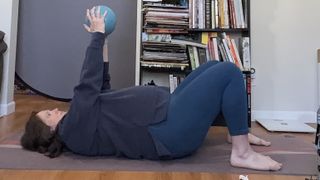 Jennifer Rizzuto performs a chest press with a Pilates ball inside on a mat. She is lying on the floor with her knees bent and feet close to her buttocks. Her arms are straight and she is holding a Pilates ball overhead.