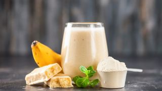 A clear glass of protein shake sits on a grey surface next to a scoop or protein powder, a green herb, a snapped chocolate bar and a banana.
