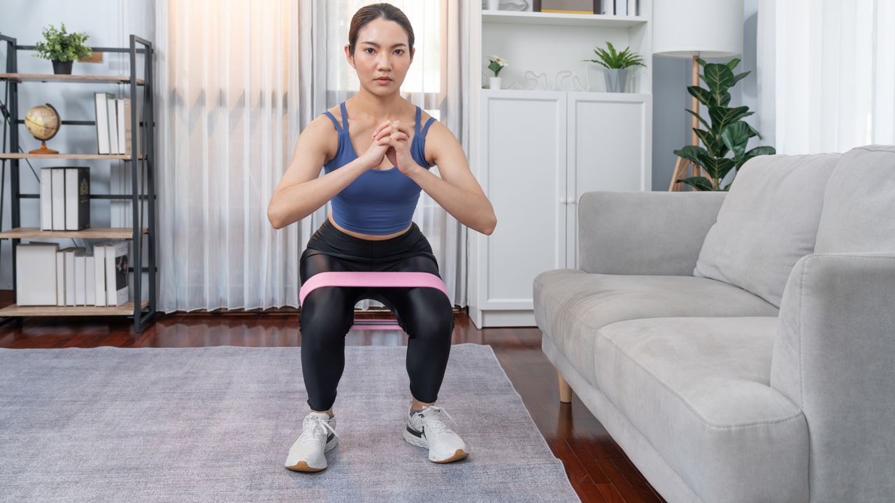 woman performing a squat with a short loop resistance band around her legs, facing the camera in a living room setting.