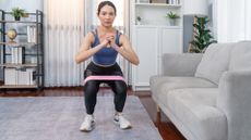 woman performing a squat with a short loop resistance band around her legs, facing the camera in a living room setting. 