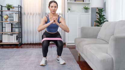 woman performing a squat with a short loop resistance band around her legs, facing the camera in a living room setting. 