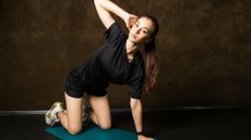A woman on a mat in a dark room performs a thoracic stretch. She is balancing on her knees and her left hand. Her right hand is behind her head, elbow bent and pointing upwards.