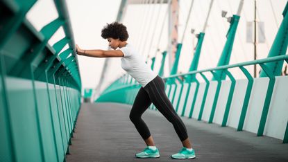 A woman stands doing a warm-up on a bridge. She is leaning against the bridge with her hands, with her left leg extended behind her and her right leg gently bent in front. 