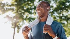 A smiling man stands outside, holding the ends of a sweat towel that is looped around his neck. He's wearing a light t-shirt and has earbuds in; he also has a sports watch strapped on his wrist.
