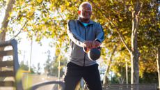 A man performs a kettlebell swing in a park. He is standing, legs straight, kettlebell grasped in his hands. His arms are also straight and the bell is swinging in front of him. Behind him we see trees and a bench.