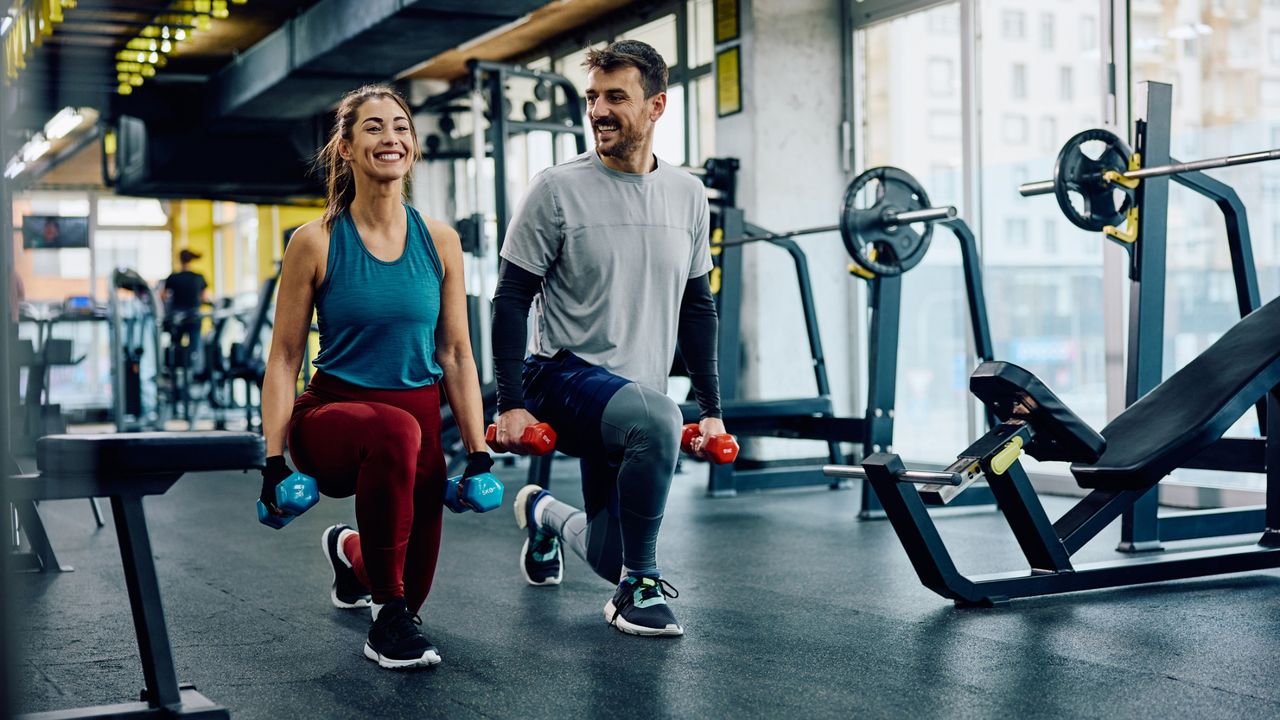 woman and man holding two dumbbells each performing a forward lunge in a gym environment. They're facing the camera and smiling.