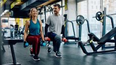 woman and man holding two dumbbells each performing a forward lunge in a gym environment. They're facing the camera and smiling. 