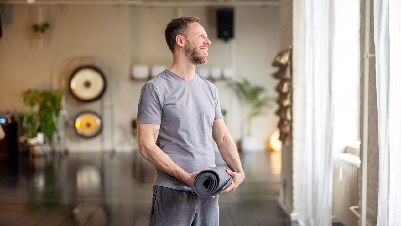 A man stands wearing loose-fitting clothing in a yoga studio, with a rolled up mat in his hands. Behind him we see plants, soft lighting and brass gongs.