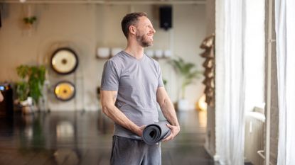 A man stands wearing loose-fitting clothing in a yoga studio, with a rolled up mat in his hands. Behind him we see plants, soft lighting and brass gongs.