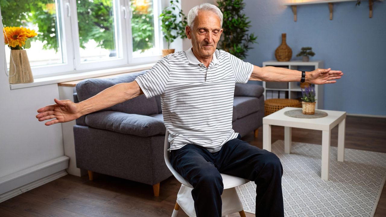 man sat on a chair in a living room setting facing the camera with his eyes closed and arms out to the sides at shoulder height.
