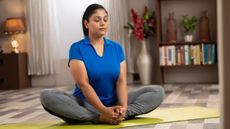 A woman practices a butterfly yoga pose at home on a mat with her eyes close. She's wearing a shirt and leggings. Her knees are bent out to the sides and her feet are touching. She sits upright and grasps both feet in her hands. Behind her we see shelves filled with books, plants and vases.