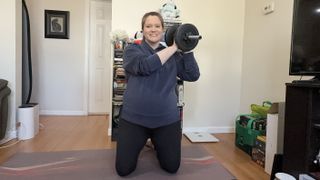 Jennifer Rizzuto performs a kneeling overhead dumbbell pass at home. She is on her knees, with her torso straight, and a dumbbell held on one shoulder. Behind her we see a bookshelf and a hallway.