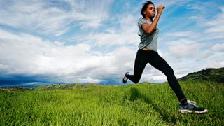 A woman runs in a hilly location over green grass with a cloud-filled blue sky behind her.