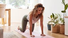 A woman is on a workout mat on her hands and knees, with her arms straight. Her eyes are closed. Behind her we see a window, table, chairs and plants.