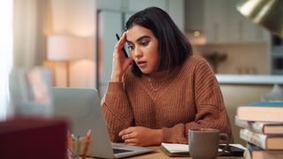 Woman sits in front of laptop at home with her fingers on her temple and a focused look on her face