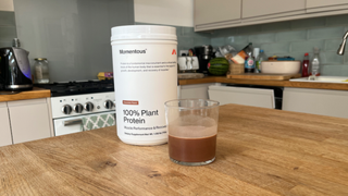 A white tub of Momentous Plant Protein powder sits next to a glass half-filled with milky brown liquid on a wooden tablet. In the background we see a kitchen sink, fruit and a kettle.