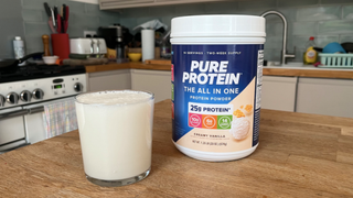 A 2lb tub of Pure Protein powder sits on a wooden table next to a glass filled with milk liquid. In the background we can see a kitchen sink, along with knives, pans and dishes.