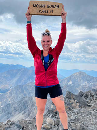 Woman in sportswear stands on top of mountain holding sign that reads "MOUNT BORAH 12,662 ft" triumphantly above her head