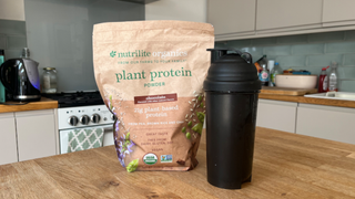 A brown bag of nutrilite organic plant protein powder sits next to a black drink flask on a wooden table. Behind this, we see a kitchen, with bottles, cleaning equipment and an oven.